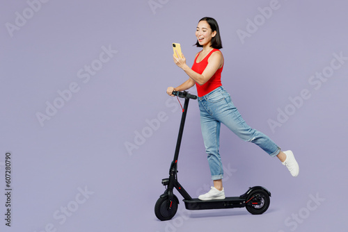 Full body sideways young woman of Asian ethnicity she wear casual clothes red tank shirt riding electric scooter use mobile cell phone isolated on plain pastel light purple background studio portrait