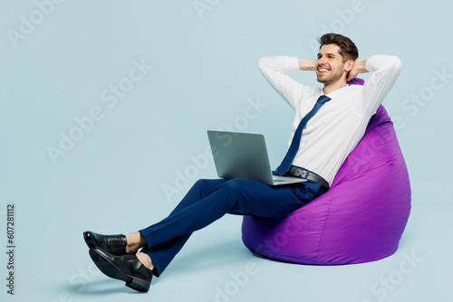 Full body young employee IT business man corporate lawyer wearing classic formal shirt tie work in office sit in bag chair hold use work on laptop pc computer isolated on plain pastel blue background.