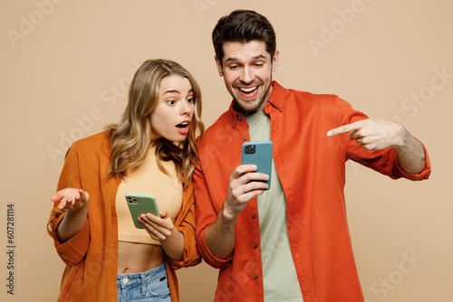 Young happy confused couple two friends family man woman wear casual clothes hold use point finger on mobile cell phone together isolated on pastel plain light beige color background studio portrait.