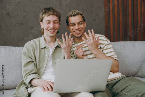 Obraz na plátně Young happy couple two gay men wear casual clothes use laptop pc tells parents about marriage proposal shows hands ring sits on sofa couch at home room indoor