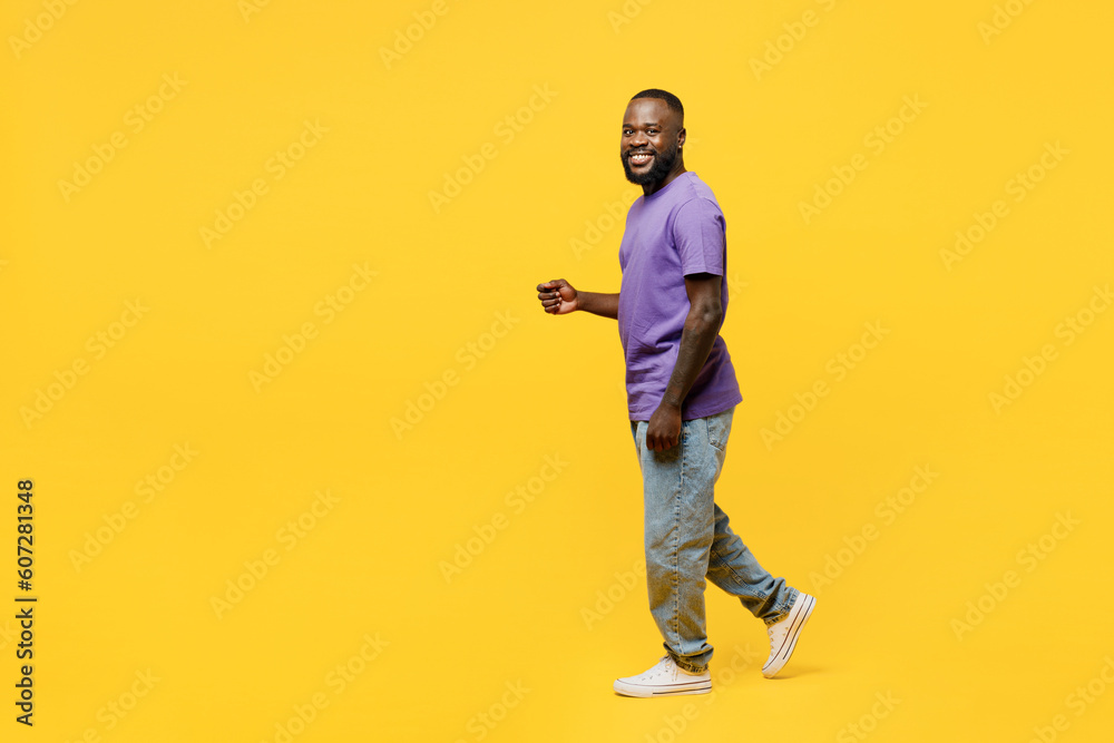 Full body side view young man of African American ethnicity he wear casual clothes purple t-shirt looking camera walk go stroll isolated on plain yellow background studio portrait. Lifestyle concept. Full body side view young man of African American ethnicity he wear casual clothes purple t-shirt looking camera walk go stroll isolated on plain yellow background studio portrait. Lifestyle concept.