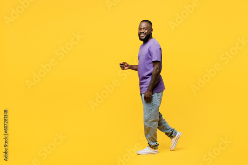 Full body side view young man of African American ethnicity he wear casual clothes purple t-shirt looking camera walk go stroll isolated on plain yellow background studio portrait. Lifestyle concept.