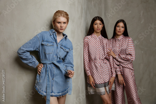 Three fashion models. Asians in identical looks with the pattern. Dress and jumpsuit. Blonde in blue denim, jeans. Beautiful young women. Sandy beige textured wall