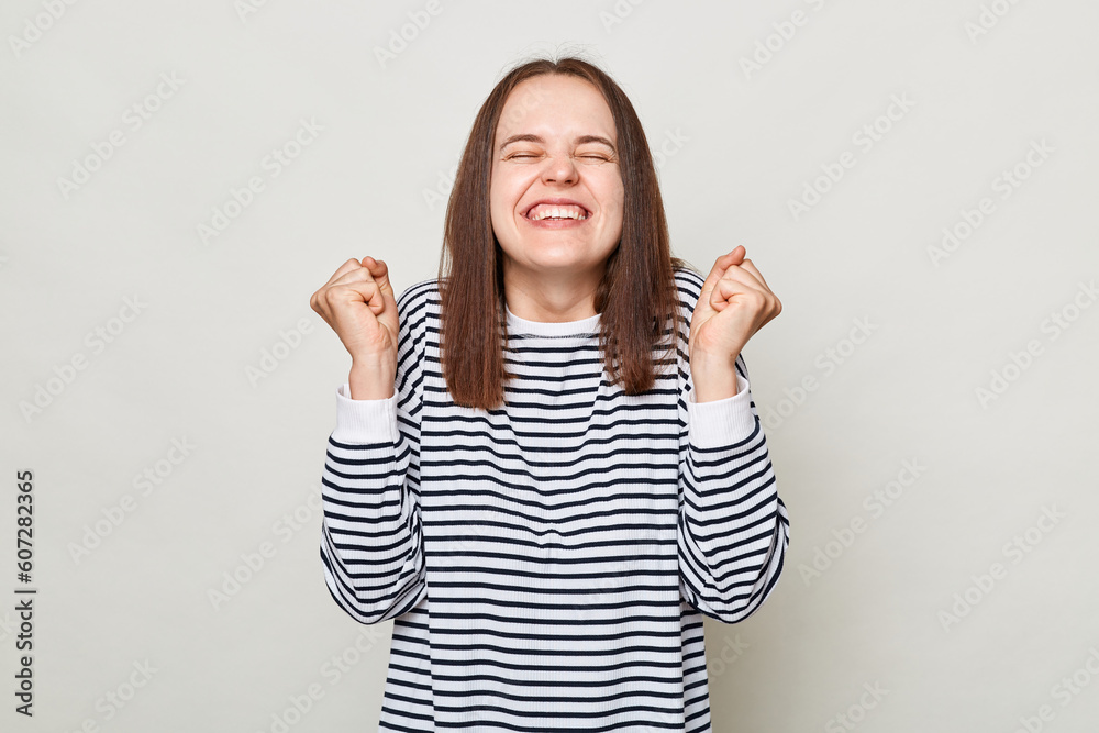 Overjoyed smiling woman with brown hair wearing striped shirt standing isolated over gray background keeps eyes closed clenched fist celebrating successful achievement. Overjoyed smiling woman with brown hair wearing striped shirt standing isolated over gray background keeps eyes closed clenched fist celebrating successful achievement.