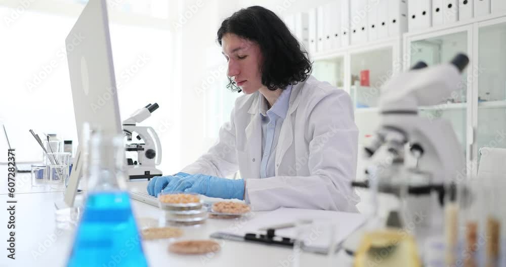 Handsome young laboratory assistant using computer working in laboratory and analyzer of cereals and grains. Research chemistry and eco products check for toxins Handsome young laboratory assistant using computer working in laboratory and analyzer of cereals and grains. Research chemistry and eco products check for toxins
