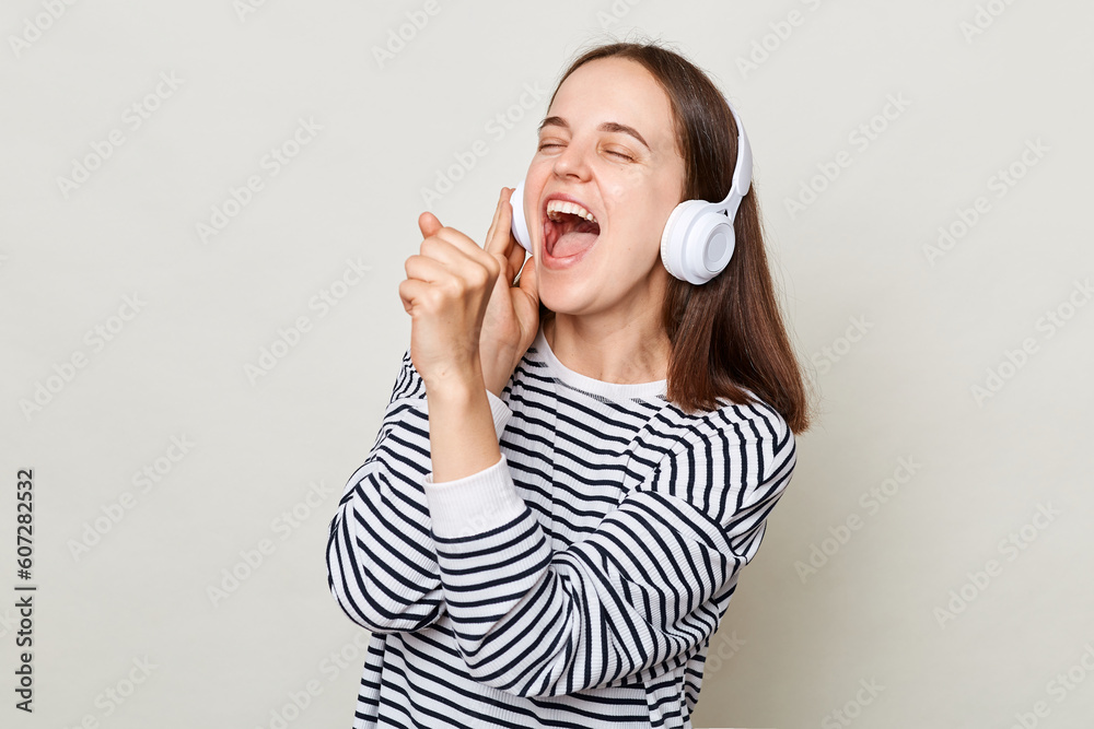 Excited festive woman with brown hair wearing striped shirt standing isolated over gray background listening to music on headphone pretending sing on microphone. Excited festive woman with brown hair wearing striped shirt standing isolated over gray background listening to music on headphone pretending sing on microphone.