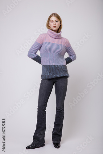 Fashion photo of beautiful elegant young woman in a pretty triple color pink, blue, gray sweater, blouse, black jeans, denim posing on white, soft gray background. Blonde.