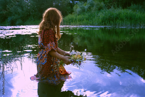 Fototapeta Naklejka Na Ścianę i Meble -  girl near river and holding flower wreath, abstract natural background. esoteric ritual for summer solstice, wiccan Litha sabbat. ceremony on Midsummer. pagan folk holiday Ivan Kupala. witchcraft