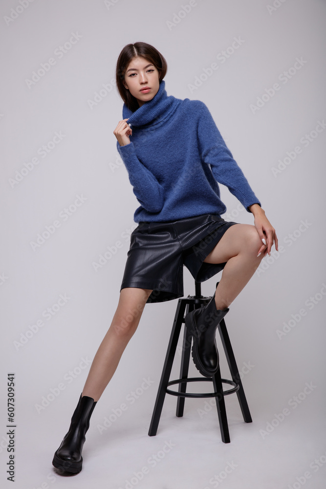 Fashion photo of a beautiful elegant young asian woman in a pretty blue sweater, blouse, black leather shorts posing on white, soft gray background. Studio Shot. Slim figure. Model sits on chair Fashion photo of a beautiful elegant young asian woman in a pretty blue sweater, blouse, black leather shorts posing on white, soft gray background. Studio Shot. Slim figure. Model sits on chair