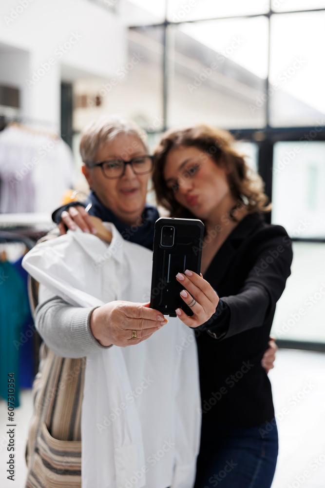 Fashionable women taking selfie with white shirt using smartphone, having fun in shopping centre. Cheerful showroom customers buying stylish clothes and merchandise in modern boutique Fashionable women taking selfie with white shirt using smartphone, having fun in shopping centre. Cheerful showroom customers buying stylish clothes and merchandise in modern boutique