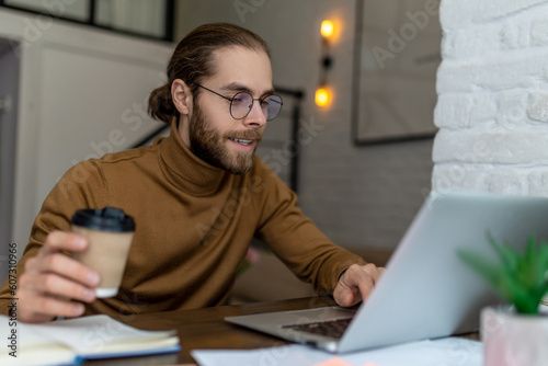 Young bearded man freelancer in glasses working at home or in cafe with laptop.