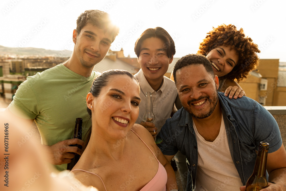 An interracial millennial group is taking a photo on a terrace while holding beers. The Caucasian girl is in the foreground picking up the smartphone. Outdoor youth party concept.