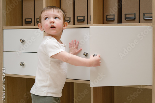 Toddler baby opens the closet door in the home living room. A small child opens a shelf drawer. Kid aged one year eight months © Андрей Журавлев