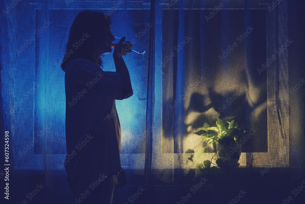 A woman drinks wine from a glass at the night dark window, low key silhouette. Problems of female alcoholism and loneliness
