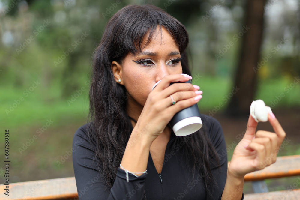 A dark-skinned woman drinks coffee and eats dessert in the park on a bench. A dark-skinned woman drinks coffee and eats dessert in the park on a bench.