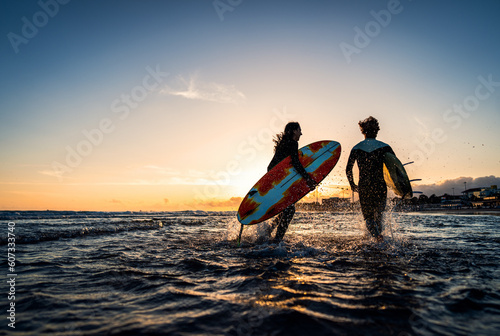 Photography Two surfers with surfboard prepares to hit the waves at sunset.