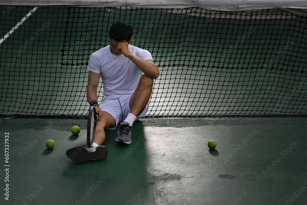 Disappointed asian male tennis player sitting on the court, upset about