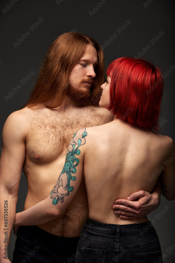 A young attractive woman with bright ruby hair, tattoos and piercings hugging a man with long hair. Half-naked lovers look into each other's eyes. Half-length studio shot, dark background. A young attractive woman with bright ruby hair, tattoos and piercings hugging a man with long hair. Half-naked lovers look into each other's eyes. Half-length studio shot, dark background.