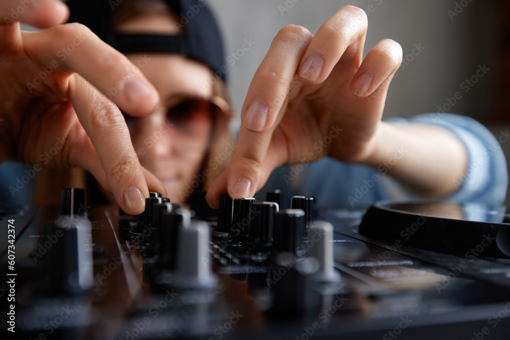 A young pretty long-haired DJ girl in a blue sweater and black baseball cap poses with a black DJ mixing console and mixes music tracks. Close-up studio shot, gray background. A young pretty long-haired DJ girl in a blue sweater and black baseball cap poses with a black DJ mixing console and mixes music tracks. Close-up studio shot, gray background.