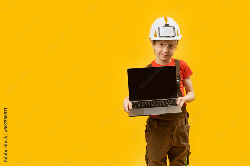 Schoolboy in work clothes and protective helmet holds laptop with empy screen. Child as architect. Choice of future profession. Copy space. Schoolboy in work clothes and protective helmet holds laptop with empy screen. Child as architect. Choice of future profession. Copy space.