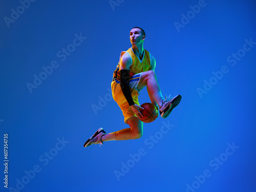 Dynamic image of professional male basketball player in motion, jumping with ball against blue studio background in neon light. Concept of professional sport, hobby, healthy lifestyle, action and