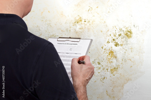 High humidity damage concept: man with a GERMAN inspection checklist in front of a white wall overgrown with mould, mildew or fungus.