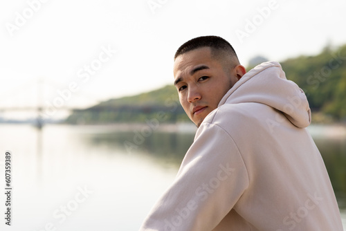 A young man stands by the river and looks at the camera over his shoulder. An Asian guy in a white hoodie poses for the camera against the backdrop of a blurred river.