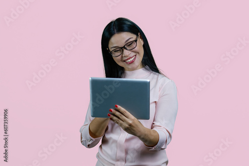 Cheerful smiling asian woman in glasses holding tablet pc. Isolated over pink background.