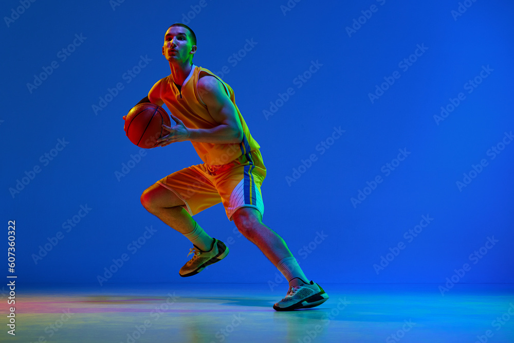 Dynamic image of young basketball player in yellow uniform during game, training against blue studio background in neon light. Concept of professional sport, hobby, healthy lifestyle, action, motion
