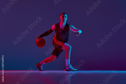 Young sportive man  basketball player in uniform during game  dribbling ball against purple studio background in neon light. Concept of professional sport  hobby  healthy lifestyle  action and motion
