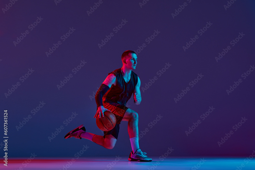 Concentrated young man, basketball player during game, training, dribbling ball against purple studio background in neon light. Concept of professional sport, hobby, healthy lifestyle, action, motion Concentrated young man, basketball player during game, training, dribbling ball against purple studio background in neon light. Concept of professional sport, hobby, healthy lifestyle, action, motion