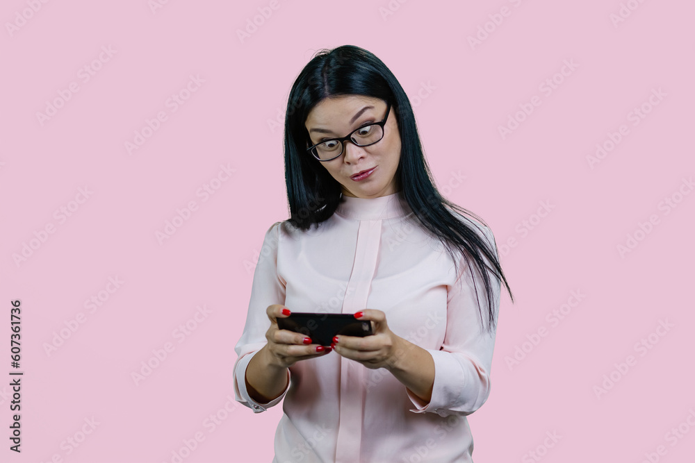 Surprised young asian woman with smartphone in her hands. To see something unexpected. Isolated over pink background.