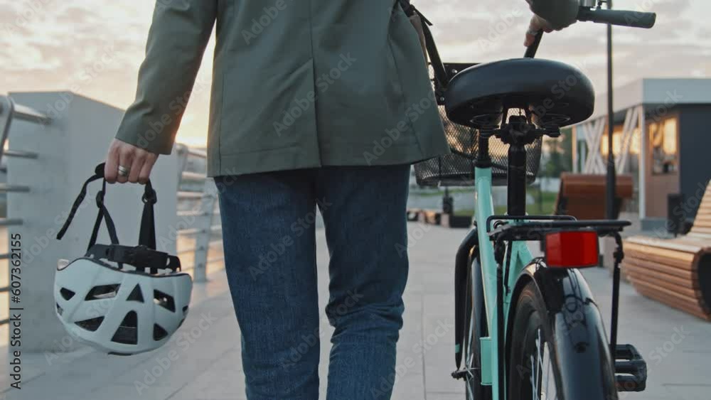 Medium section rear view slow motion of unrecognizable woman with bicycle holding helmet walking along river bank in morning Medium section rear view slow motion of unrecognizable woman with bicycle holding helmet walking along river bank in morning