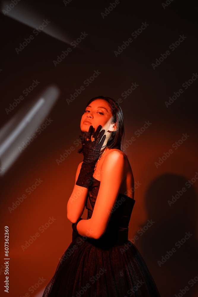 attractive asian woman with wet hairstyle posing in strapless dress with tulle skirt and black gloves with rings while standing on dark orange background with red lighting, model, looking at camera