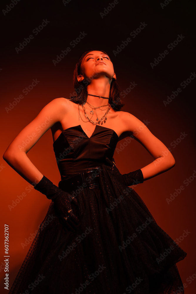 stylish asian woman with closed eyes and wet hairstyle posing with hands on hips in black strapless dress with tulle skirt and gloves while standing on orange background with red lighting, young model