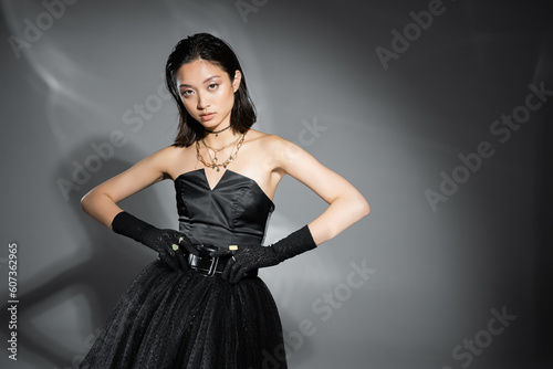 charming asian young woman with short hair posing in black strapless dress with tulle skirt touching belt and looking at camera on grey background, wet hairstyle, golden jewelry