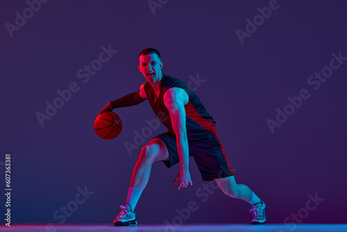Young muscular man, basketball player in motion, dribbling ball against purple studio background in neon light. Winner. Concept of professional sport, hobby, healthy lifestyle, action and motion