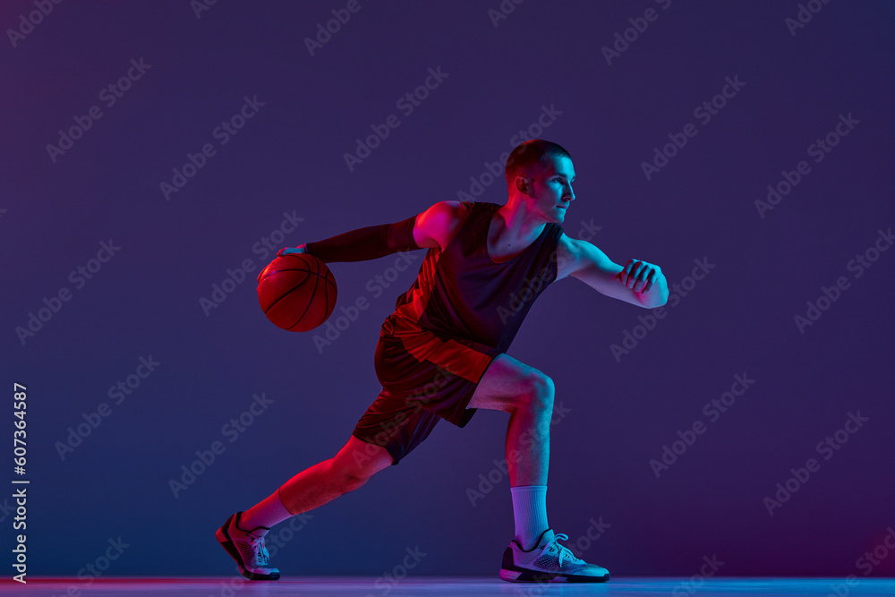 Young athletic man, basketball player in motion with ball against purple studio background in neon light. Champion. Concept of professional sport, hobby, healthy lifestyle, action and motion Young athletic man, basketball player in motion with ball against purple studio background in neon light. Champion. Concept of professional sport, hobby, healthy lifestyle, action and motion