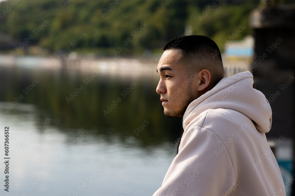 A young man stands on the embankment and admires the view of the river. An Asian man in a white hoodie stands in front of a blurred river. A young man stands on the embankment and admires the view of the river. An Asian man in a white hoodie stands in front of a blurred river.