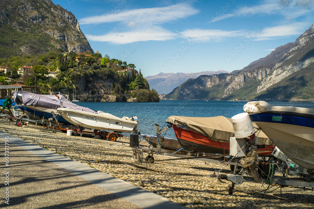 Buildings of the city of Lecco on the shores of Lake Como surrounded by mountains in the background. Sailboats on the waters of the lake and the waterfront.