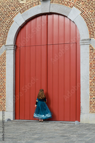 A little girl in a medieval dress is knocking on the big red gates of an ancient castle. vertical format