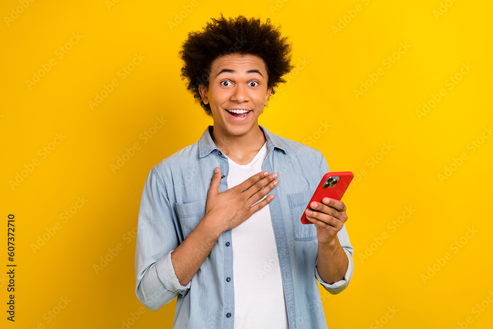 Photo of impressed man dressed denim shirt hold smartphone hand on chest staring at unbelievable sale isolated on yellow color background