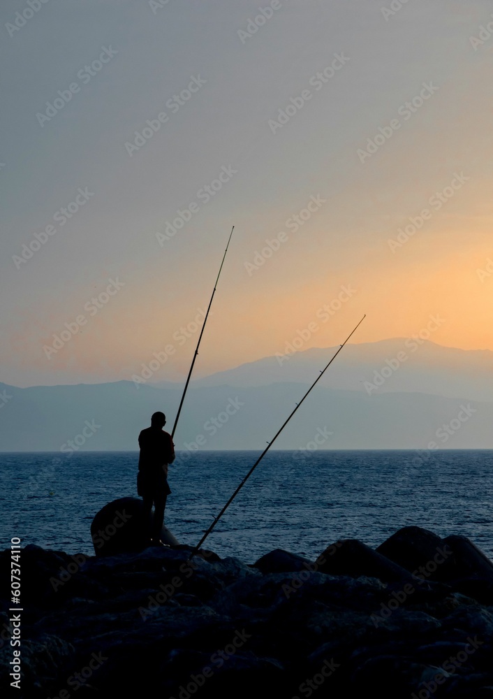 Silhouette of fisherman standing on the shore with rod on sunset sky background