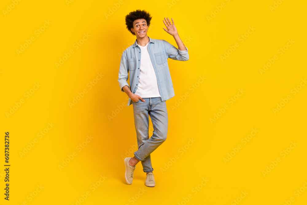 Full length photo of optimistic cool man dressed denim shirt trousers arm in pocket waving palm say hi isolated on yellow color background Full length photo of optimistic cool man dressed denim shirt trousers arm in pocket waving palm say hi isolated on yellow color background