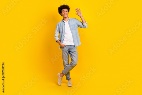 Full length photo of optimistic cool man dressed denim shirt trousers arm in pocket waving palm say hi isolated on yellow color background