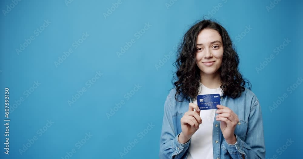 Happy young brunette with credit card. Static shot of young female bank client looking at camera with smile and shaking credit card against blue background Happy young brunette with credit card. Static shot of young female bank client looking at camera with smile and shaking credit card against blue background