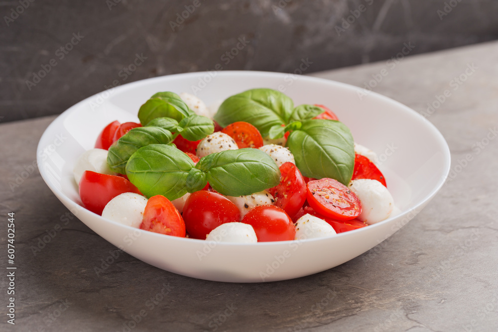 Caprese salad with tomato, mozzarella, basil and olive oil on white plate on dark table. Isometric style. Mediterranean cuisine. Caprese salad with tomato, mozzarella, basil and olive oil on white plate on dark table. Isometric style. Mediterranean cuisine.