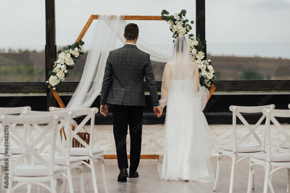 Bride and groom in a white wedding long dress walks to the wooden arch, back view. Wedding concept.