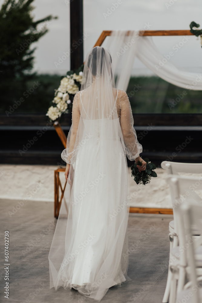 Gorgeous bride in white elegant long dress is walking tho wooden arch. Back view.