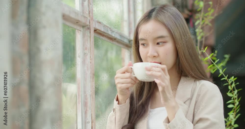 Beautiful asian woman drinking coffee enjoy flavor at trendy cafe in a rainy day. Attractive young female hold a mug with cappuccino looking outside the window and thinking. calmness, city lifestyle. Beautiful asian woman drinking coffee enjoy flavor at trendy cafe in a rainy day. Attractive young female hold a mug with cappuccino looking outside the window and thinking. calmness, city lifestyle.